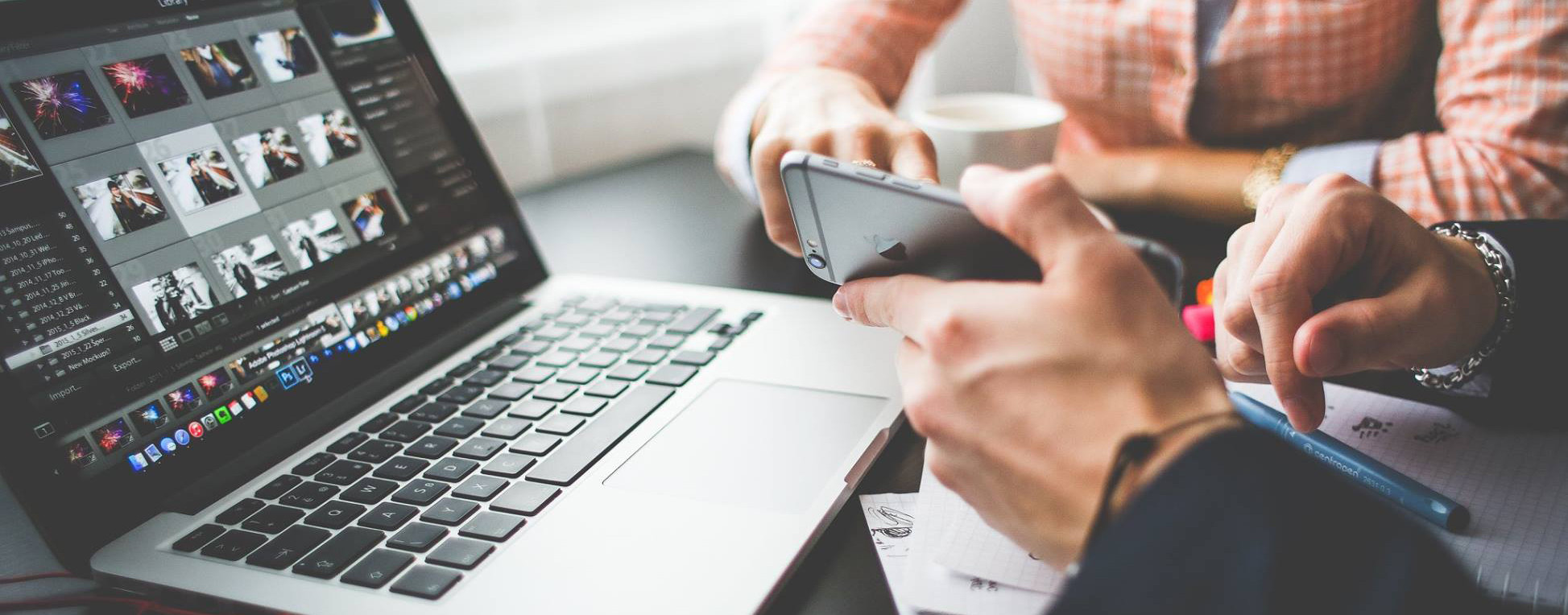 Person holding a cell phone sitting in front of a laptop showing multiple video thumbnails while another person with a beverage cup sits nearby